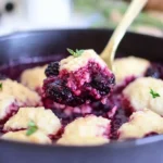 A skillet of blackberry dumplings with golden dough and vibrant blackberry sauce, served fresh and garnished with a sprig of thyme.