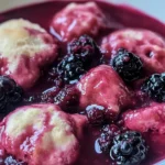 A close-up view of soft, fluffy dumplings surrounded by thick blackberry sauce and fresh blackberries.