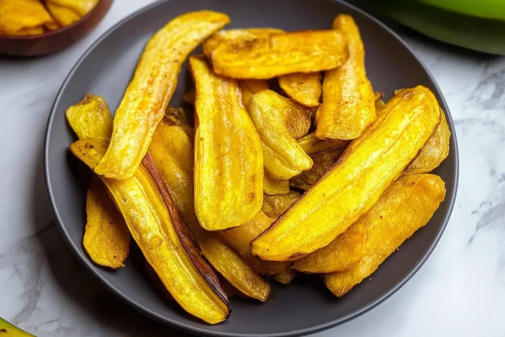 A plate of long-sliced, crispy plantain chips, golden yellow in color, arranged neatly with fresh green plantains in the background.